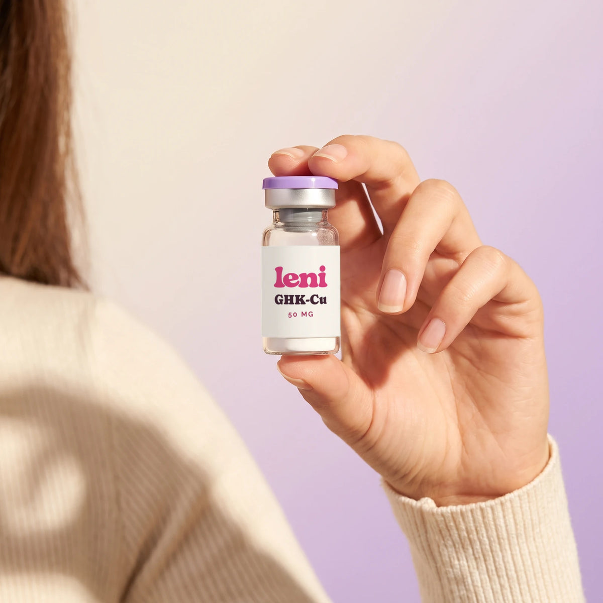 A relaxed hand holding a Leni GHK-Cu vial against a soft cream and lilac backdrop.